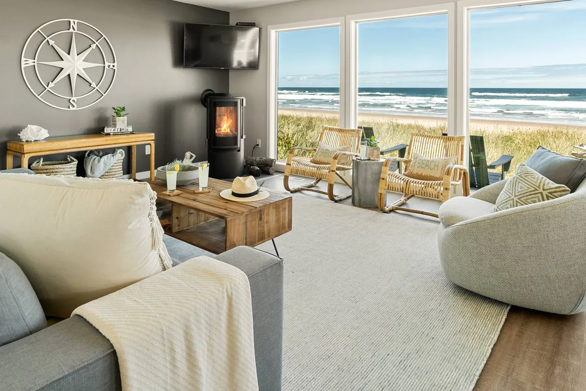 A living room interior with a modern fireplace, comfortable seating, and large windows showing an ocean view. The compass wall art adds a nautical touch while the fireplace and seating area suggest warmth and comfort for winter evenings.