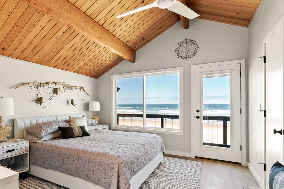 A serene bedroom featuring a bed with textured bedding, large window framing an ocean view, and exposed wooden ceiling beams. The room is bright with natural light, evoking a tranquil, coastal atmosphere ideal for relaxation.