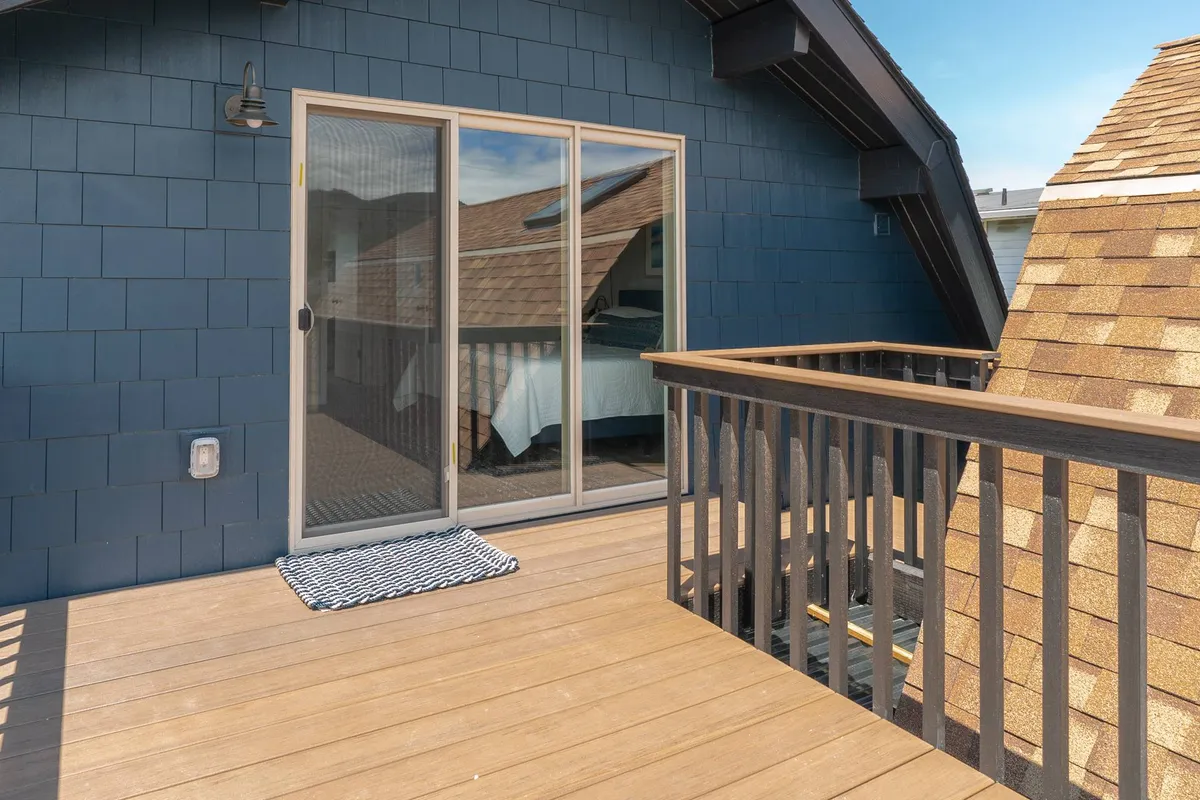 This image shows a serene exterior view of a deck area with a sliding glass door, deck railing, and a textured wall. The space appears tranquil and inviting, suggesting a peaceful spot for guests and their pets to relax outdoors after a beach outing. Early morning light and fresh air add to the welcoming atmosphere.