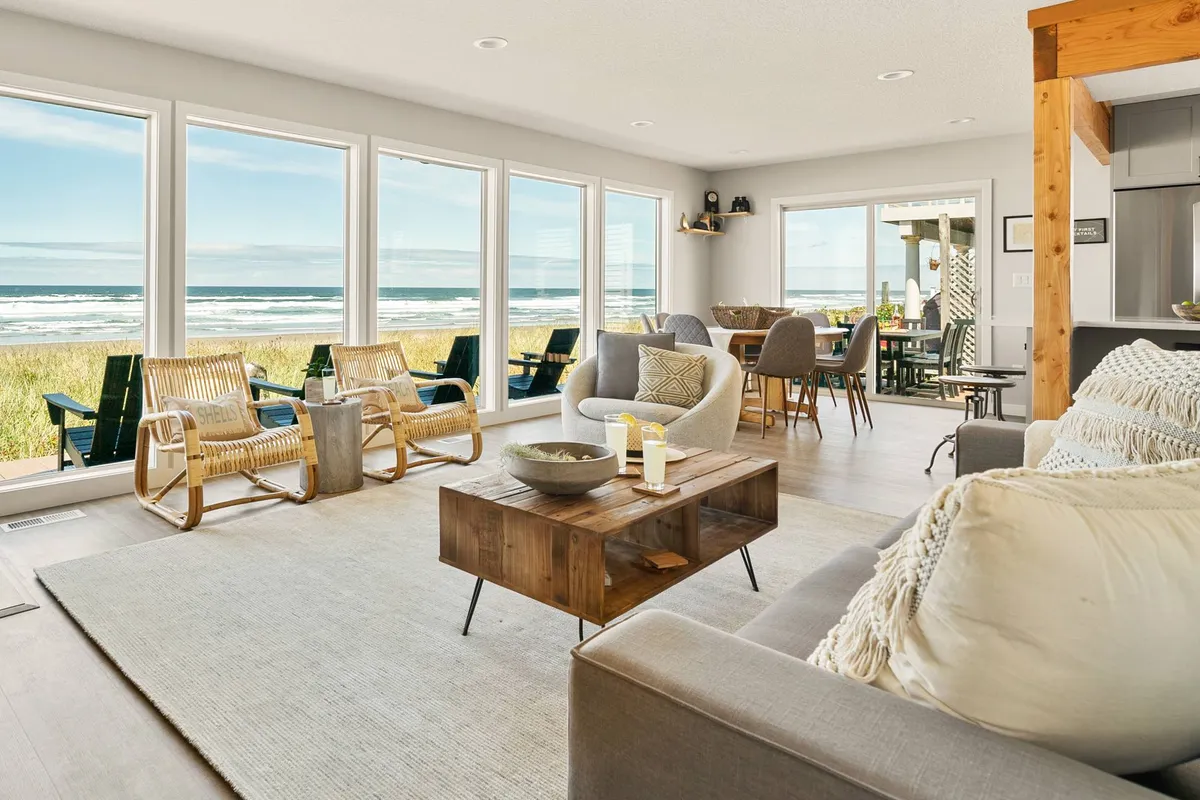 The image showcases a beautifully furnished living area with large windows framing sweeping ocean views. A cozy seating arrangement invites relaxation, while a wooden coffee table and a stylish dining table in the background hint at shared meals with loved ones. The space is bright, airy, and welcoming, seamlessly blending comfort with the stunning coastal scenery.