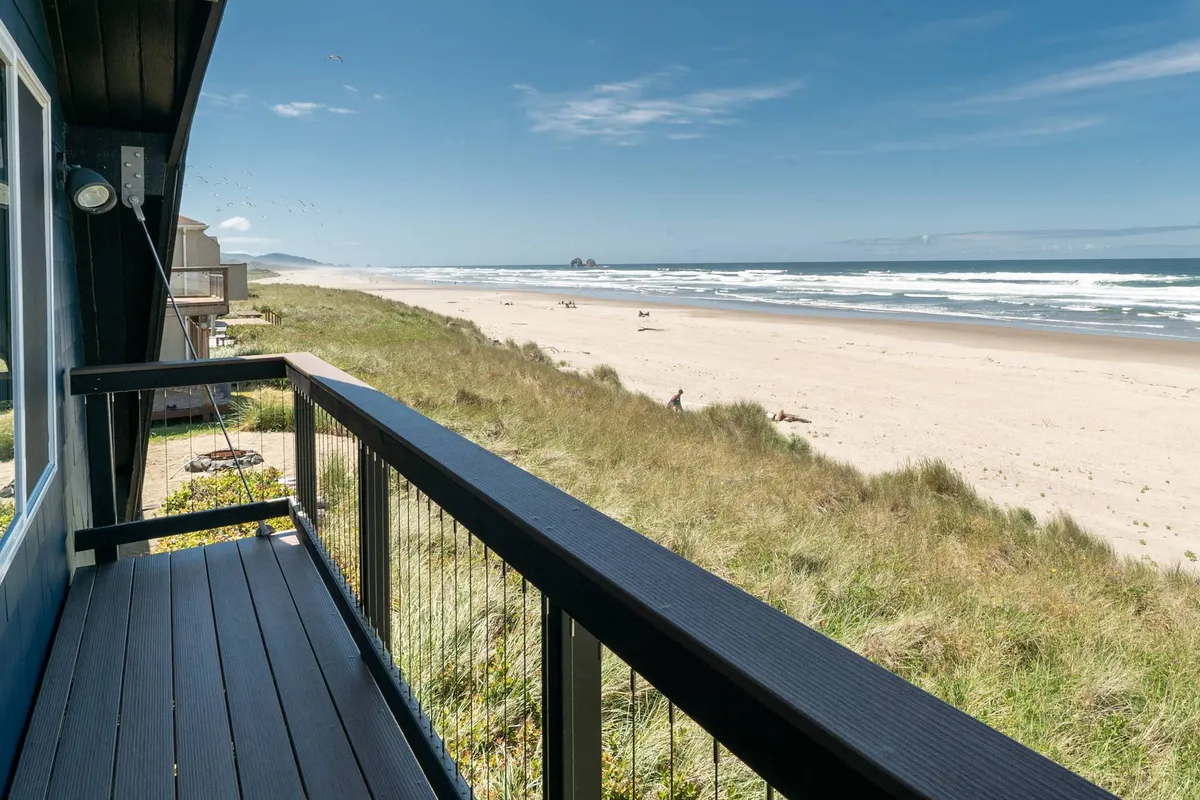 A sweeping view of Rockaway Beach’s sandy shoreline, rolling dunes, and the Pacific Ocean, framed by the deck railing in the foreground. The image captures the direct proximity of the beach to the property and hints at the inviting outdoor living space.