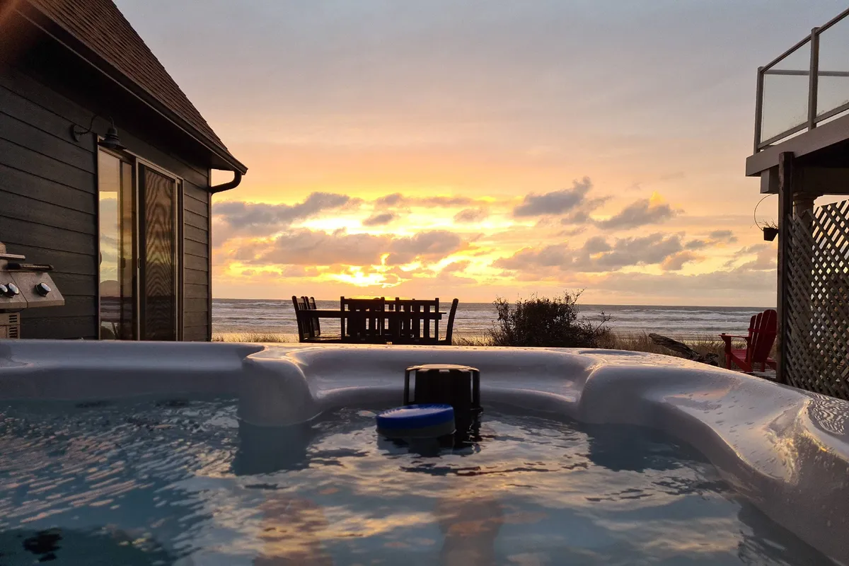 This image captures an exterior view with a private hot tub overlooking the ocean at sunset. Outdoor furniture and a dining area suggest year-round use, with the warm glow of sunset highlighting the relaxing atmosphere.