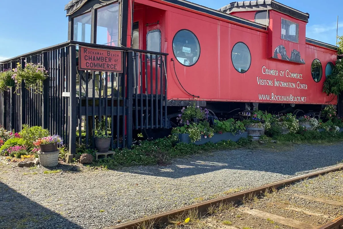 A vibrant red caboose sits in front of the Chamber of Commerce, surrounded by seasonal flower pots and informative signage. The friendly, inviting setting suggests a welcoming gateway for visitors to access information and discover hidden gems in the region.