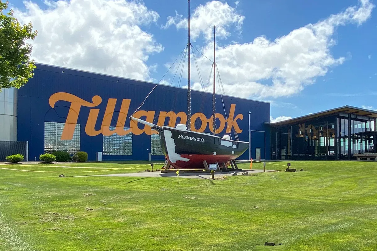 This image features the iconic Tillamook factory building with its prominent blue exterior, a boat named 'Morning Star,' and glass-walled architectural elements. Visitors can be seen enjoying the outdoor seating area, giving a sense of the lively and community-oriented atmosphere.