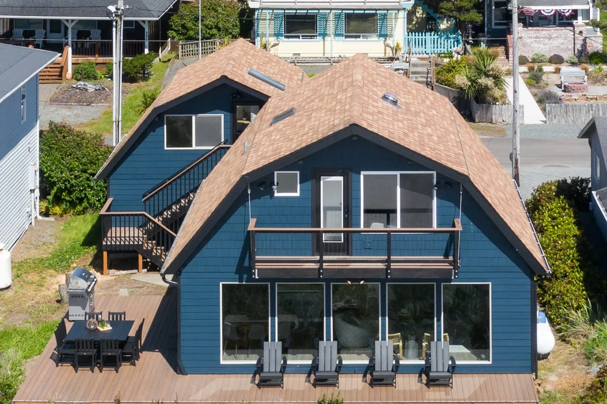 This exterior image shows a large deck with seating, grill setup, and the home’s dual A-frame structure with expansive windows. It suggests lively outdoor gatherings, barbecues, and sunny lounging.