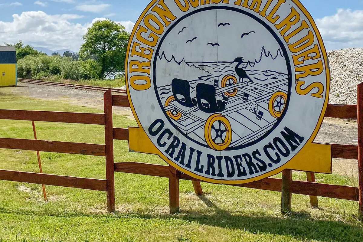 This exterior image features the Oregon Coast Trail Riders sign, railroad tracks, green grass, and a cheerful yellow building. The setting conveys a sense of outdoor adventure and connection to the local landscape, suggesting nearby pet-friendly exploration opportunities.