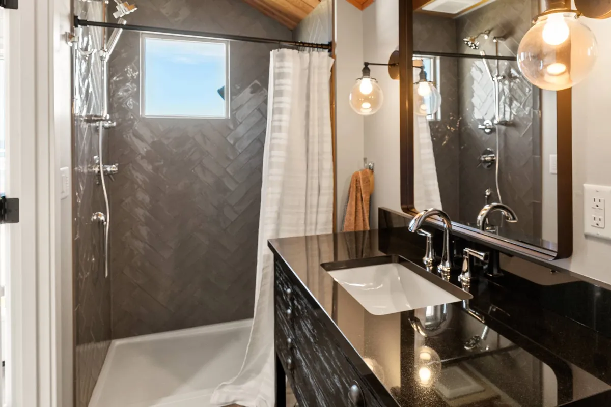 A modern bathroom with a black granite countertop, large mirror illuminated by sleek fixtures, and a stylish shower with herringbone tile. The ambiance is clean, luxurious, and inviting.