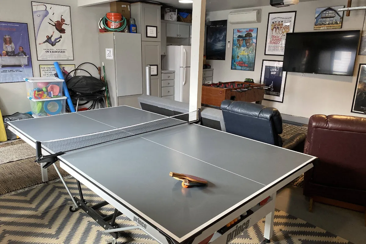 A lively game room interior featuring a ping pong table, comfortable sofa chairs, framed posters, and a large television. The space is set up for friendly competition, movie nights, and group relaxation.