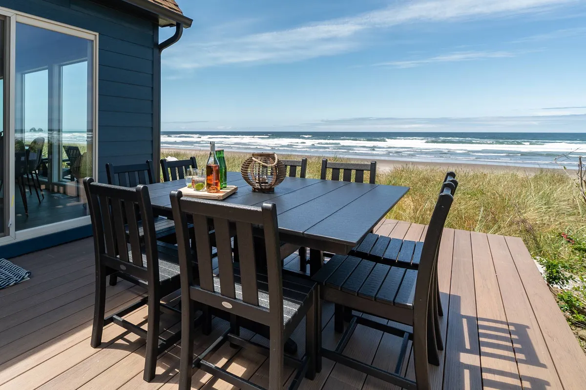 A spacious deck with an outdoor dining table set for a meal, overlooking the ocean. The table is arranged for a gathering, and the expansive view shows sunlit waves in the background, evoking a welcoming setting for group meals and sunset celebrations.