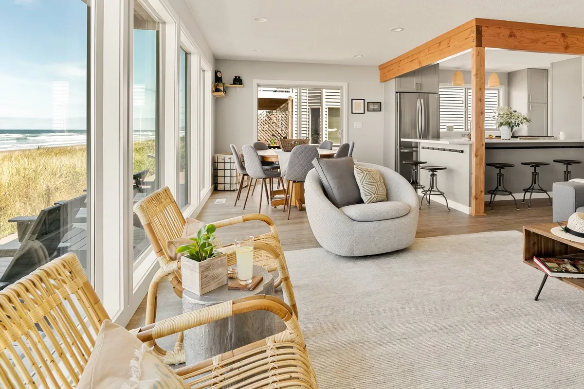 An open-concept interior space featuring a cozy seating area, dining table, and kitchen, all framed by expansive windows that overlook the beach and ocean. The space is filled with natural light, highlighting the seamless flow between the kitchen, dining, and living areas, and immediately draws the eye toward the panoramic ocean view.